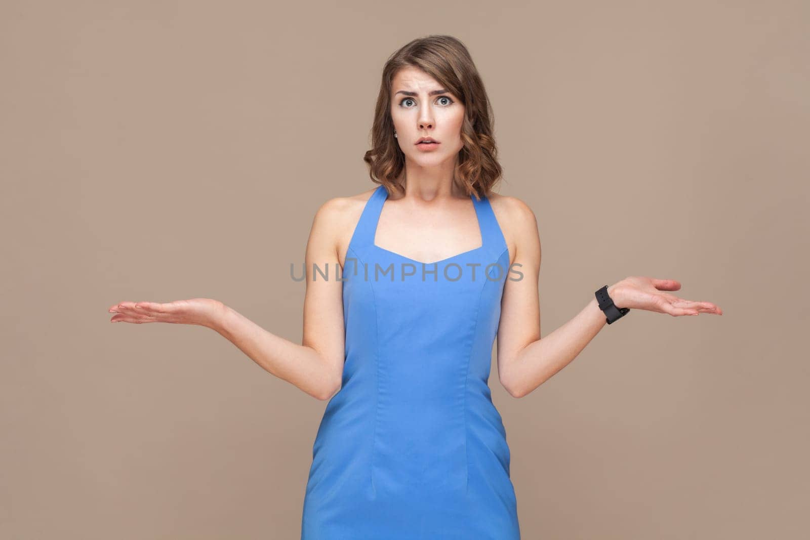 Portrait of puzzled confused young adult woman with wavy hair standing shrugging shoulders, don't know what to do, wearing blue dress. Indoor studio shot isolated on light brown background.