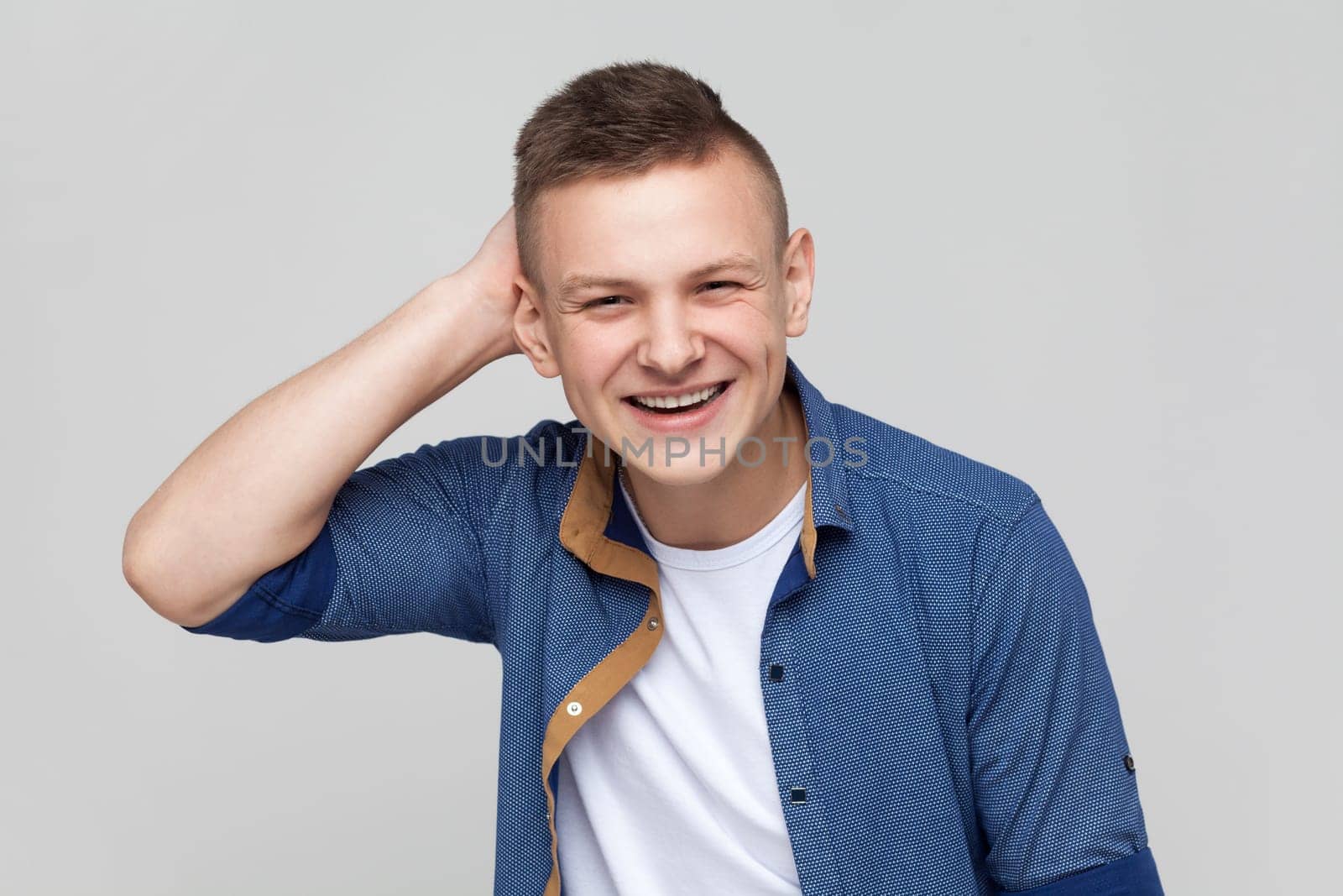 Portrait of young handsome positive friendly teenager boy wearing blue shirt looking at camera with smile, keeping hand on his head. Indoor studio shot isolated on gray background.