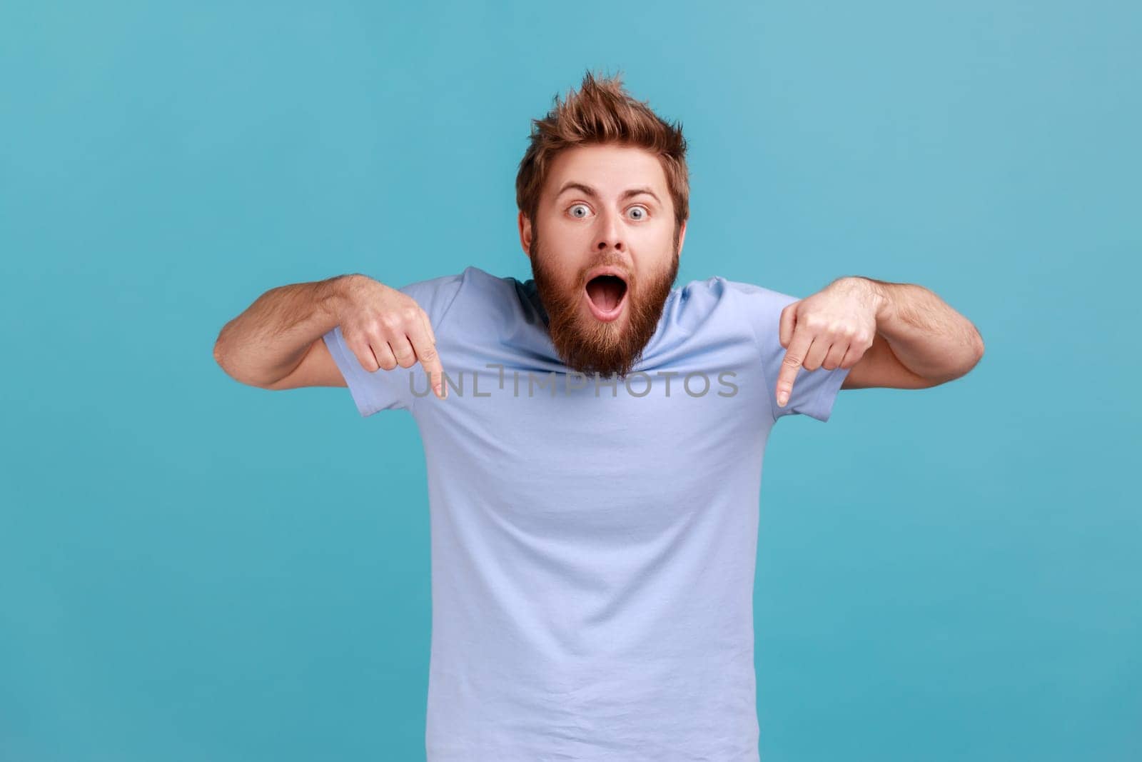 Portrait of astonished amazed bearded man pointing down and looking at camera, showing place for idea presentation, expressing surprise. Indoor studio shot isolated on blue background.