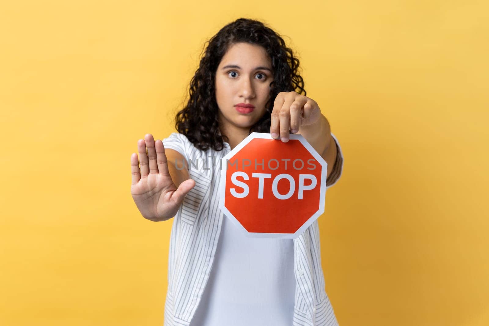 Portrait of strict woman with dark wavy hair holding red stop sign looking at camera with ban palm gesture, has strict expression, prohibition. Indoor studio shot isolated on yellow background.