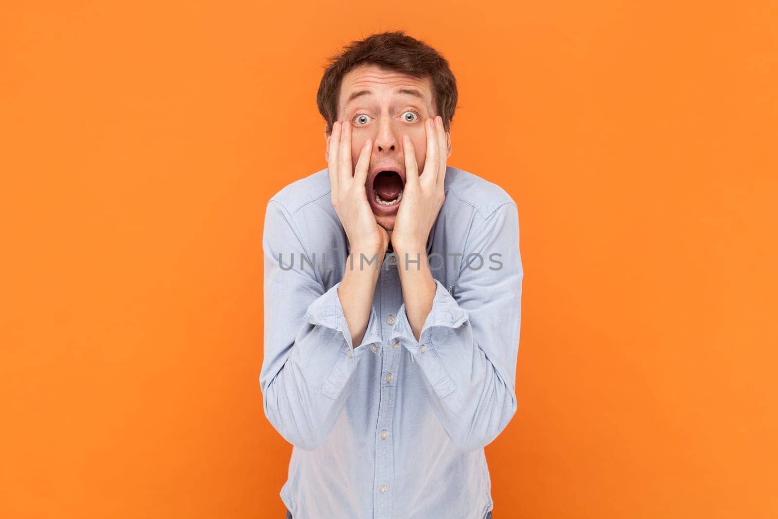 Portrait of shocked scared man looking at camera with open mouth, keeps hands on cheeks, being frighten, wearing light blue shirt. Indoor studio shot isolated on orange background.