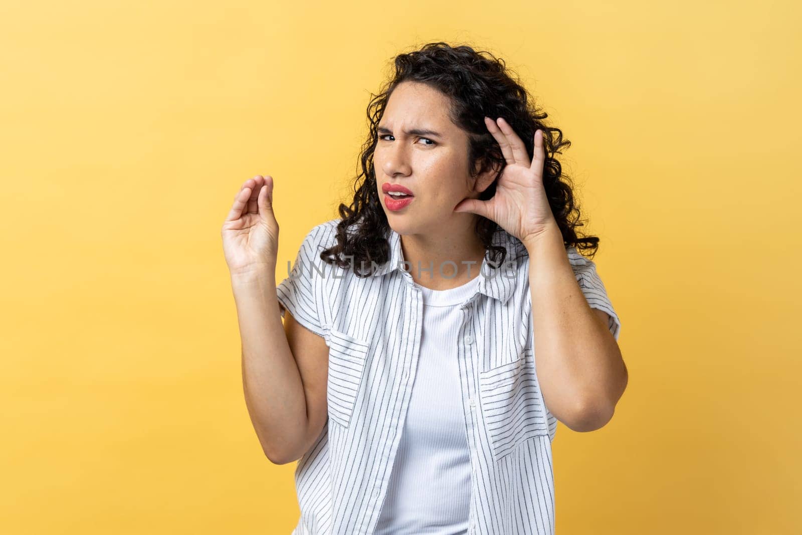 Portrait of woman with dark wavy hair holding hand near ear, listening attentively with interest private conversation, confidential talk. Indoor studio shot isolated on yellow background.