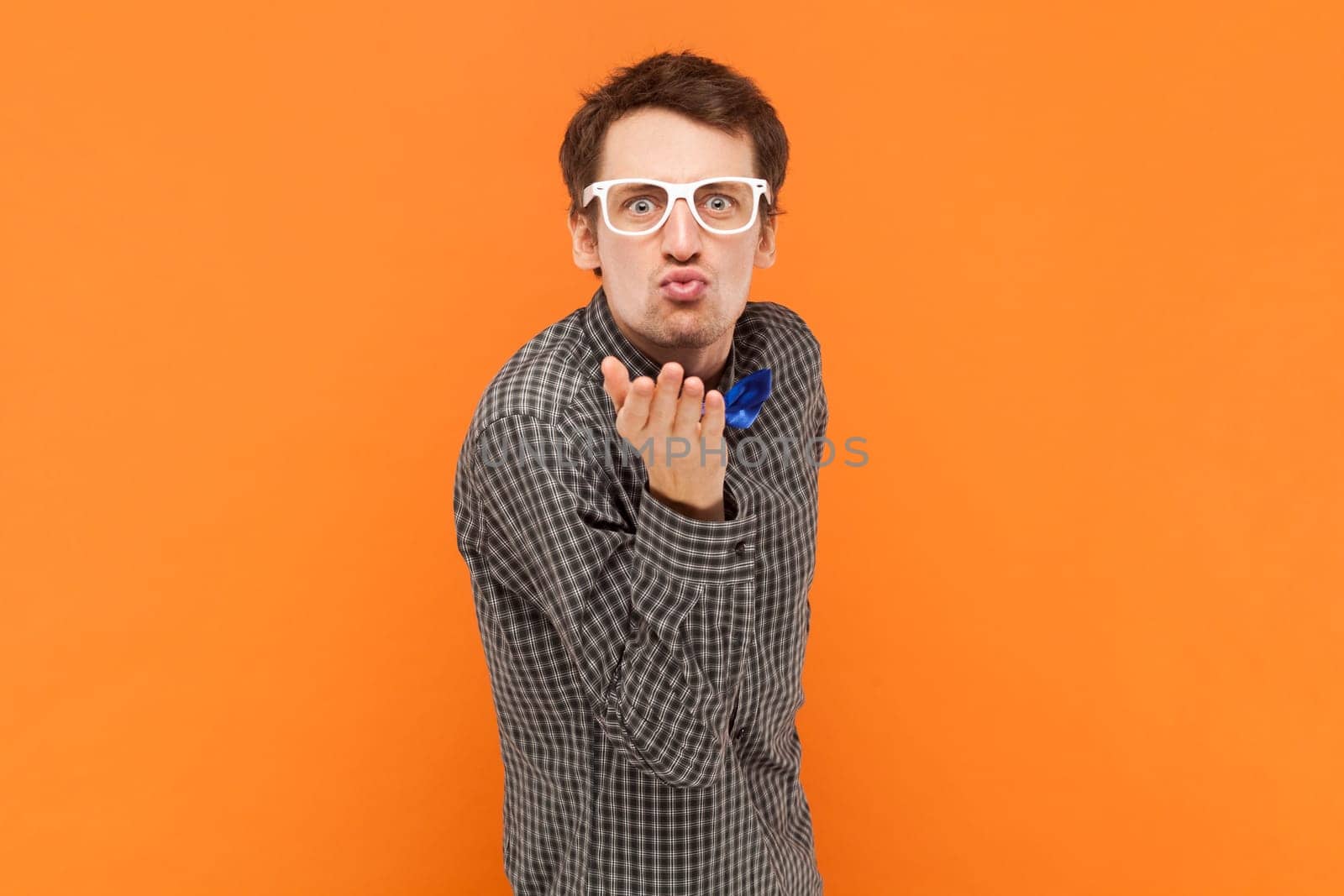 Portrait of funny positive man nerd sending air kissing, falling in love, flirting, wearing shirt with blue bow tie and white glasses. Indoor studio shot isolated on orange background.