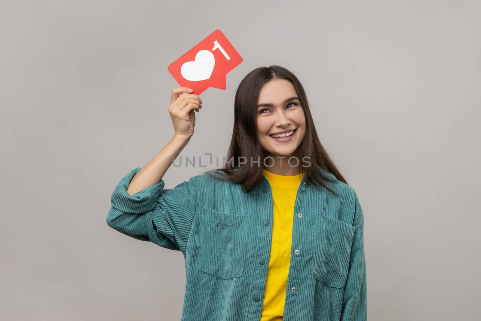 Portrait of smiling young woman holding heart icon over head, looking at camera, share blog with trendy content, wearing casual style jacket. Indoor studio shot isolated on gray background.