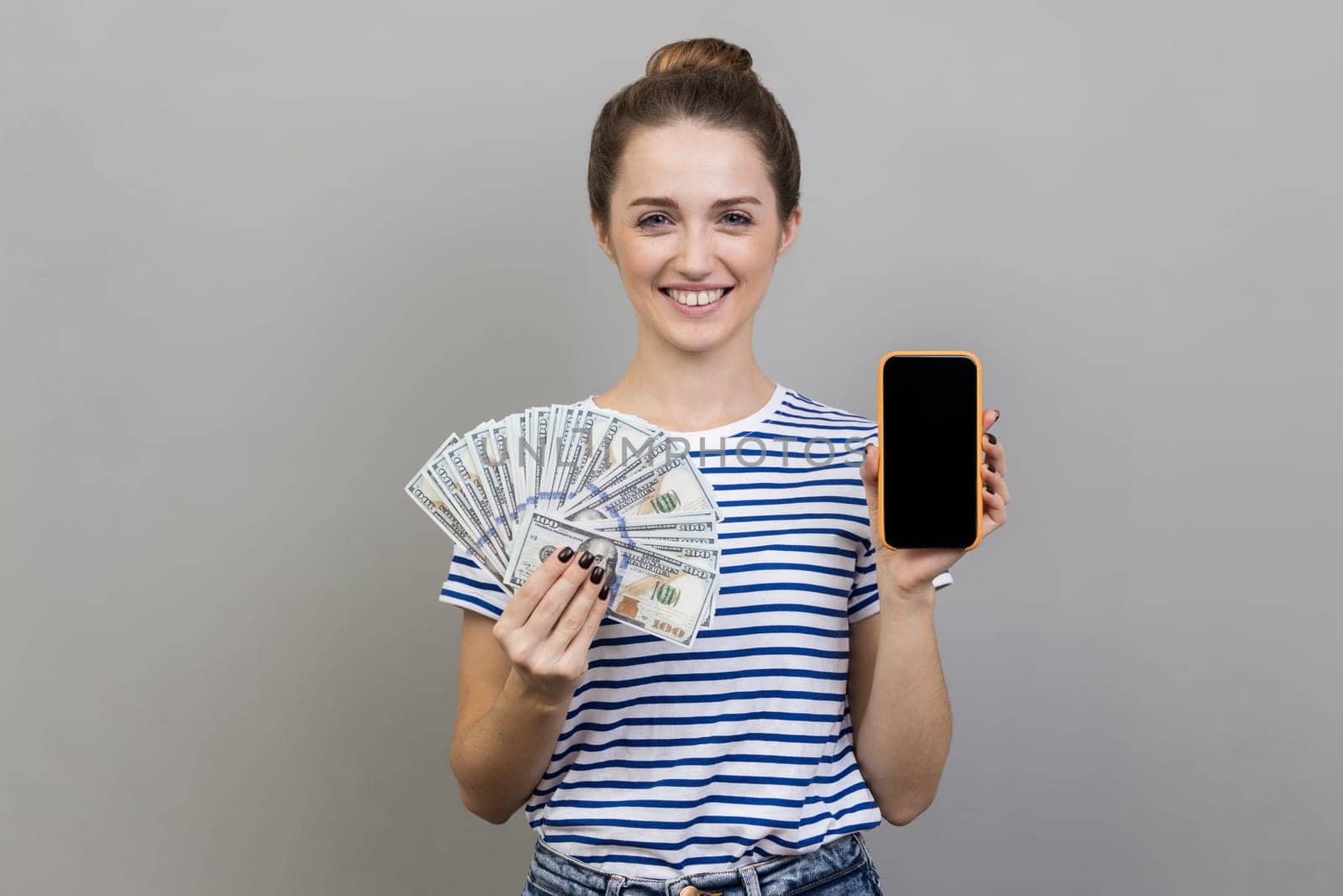 Portrait of delighted satisfied woman wearing striped T-shirt holding in hands smart phone with blank screen and dollars banknotes. Indoor studio shot isolated on gray background.