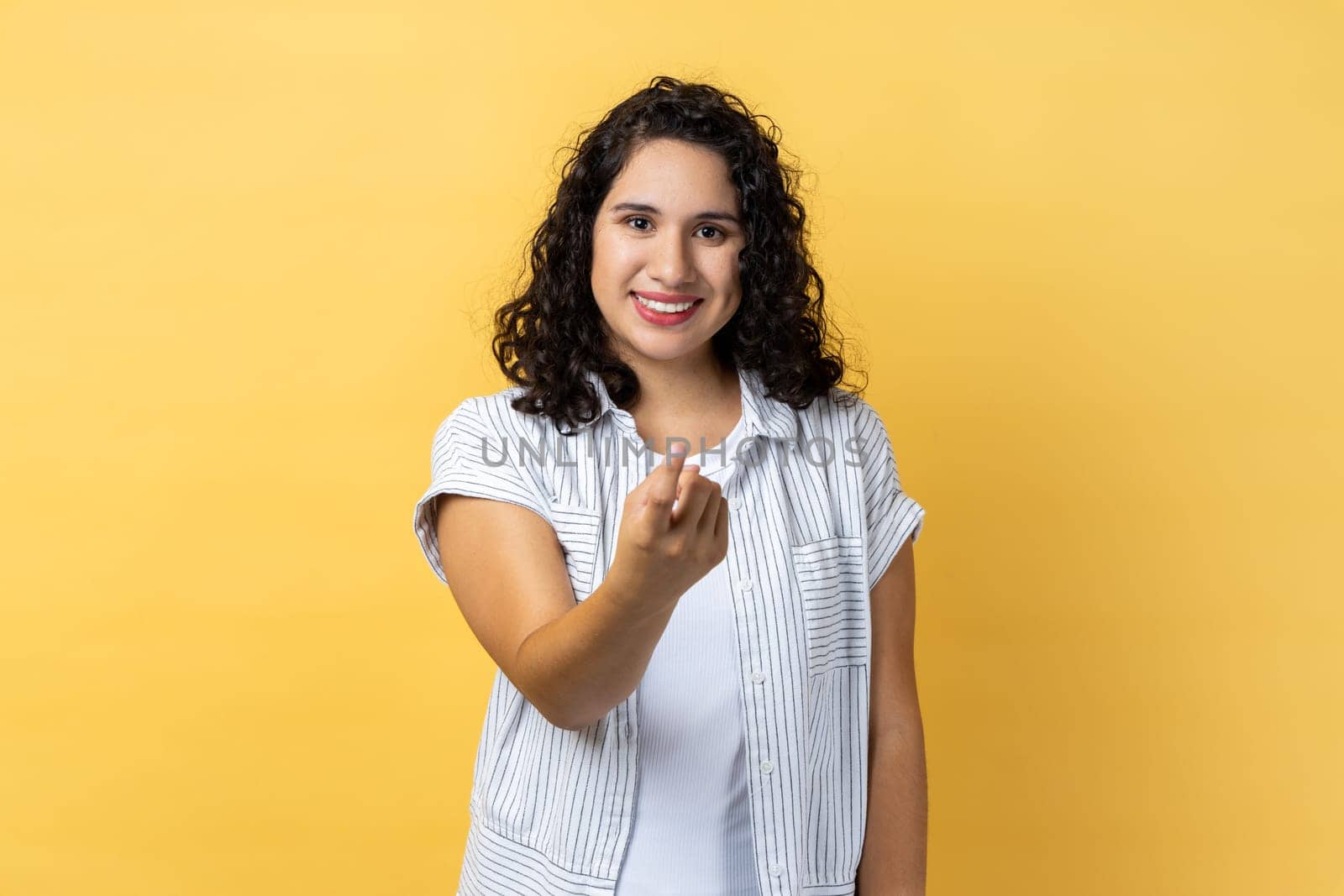 Come to me. Portrait of beautiful attractive woman with dark wavy hair making beckoning gesture and looking at camera with alluring eyes. Indoor studio shot isolated on yellow background.