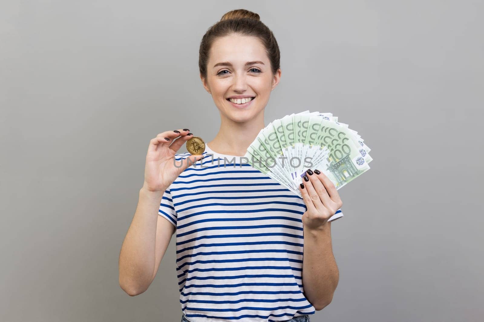 Portrait of delighted positive woman wearing striped T-shirt holding golden coin of crypto currency and big fan of euro banknotes. Indoor studio shot isolated on gray background.