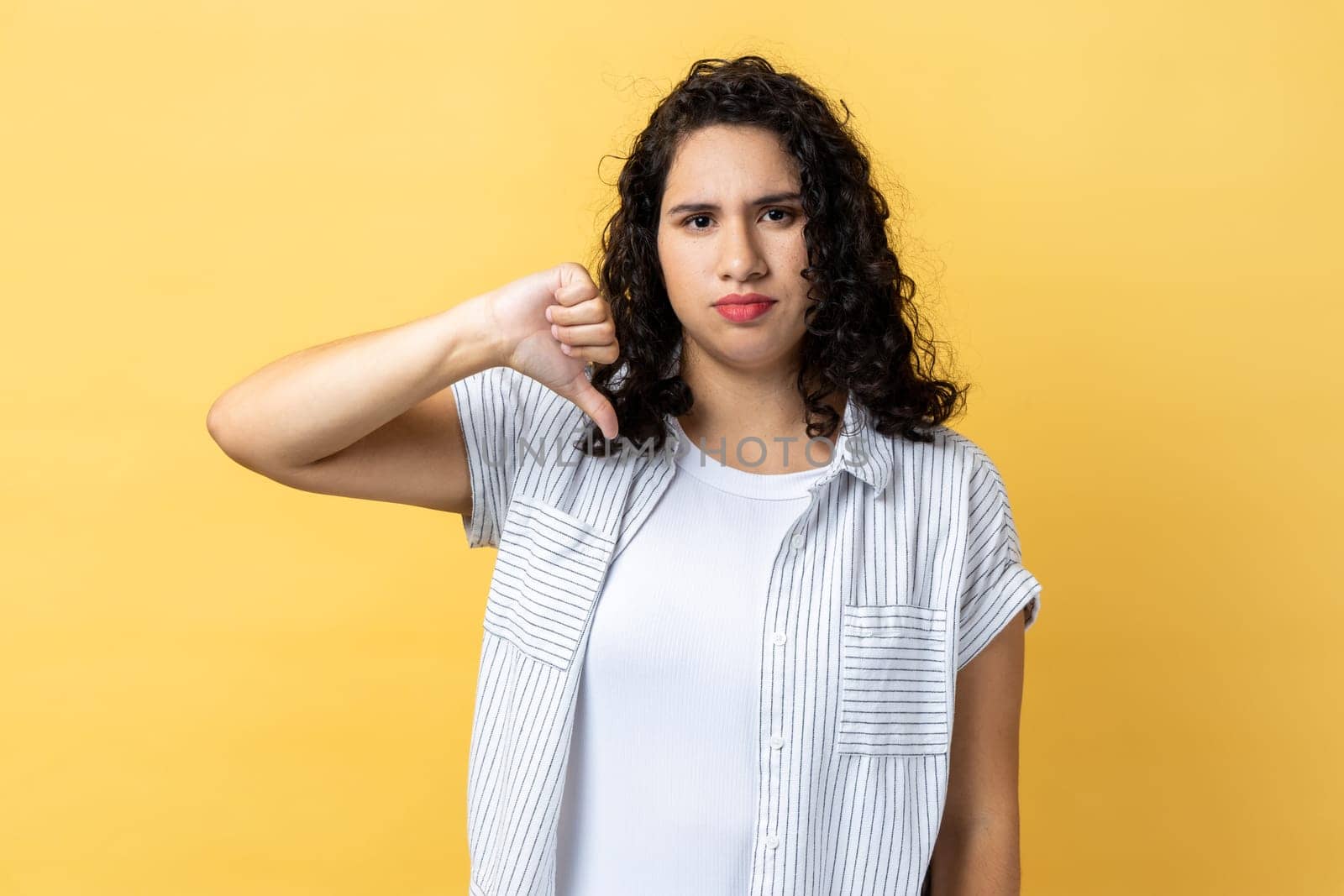 Portrait of woman with dark wavy hair criticizing bad quality with thumbs down displeased grimace, showing dislike gesture, expressing disapproval. Indoor studio shot isolated on yellow background.