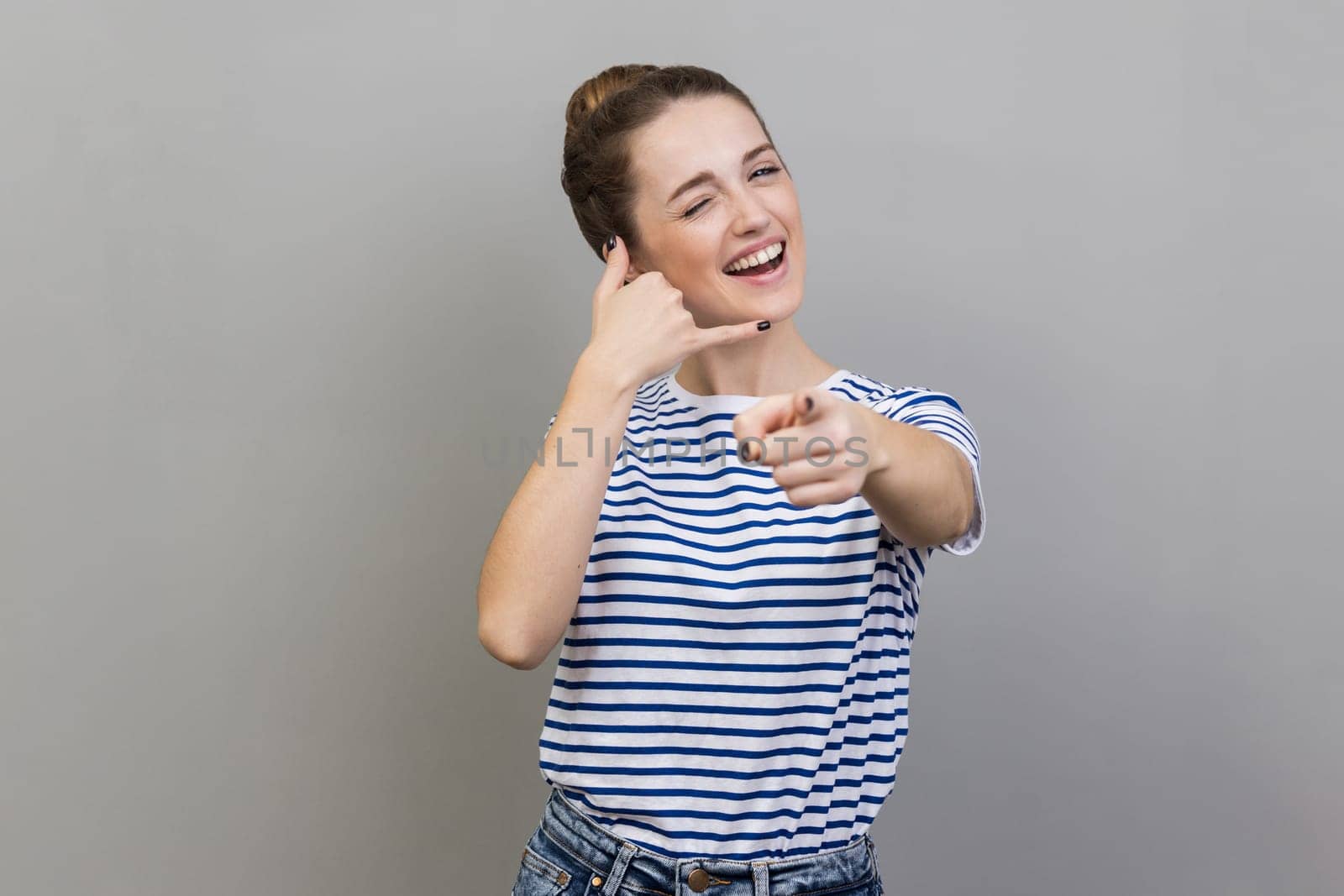 Portrait of funny beautiful woman wearing striped T-shirt standing with call gesture, looking, winking and pointing at camera, flirting. Indoor studio shot isolated on gray background.