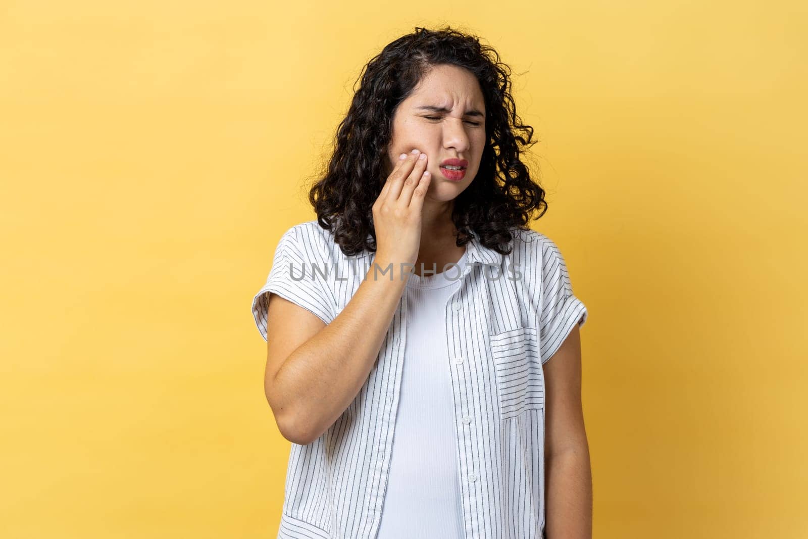 Portrait of unhealthy woman with dark wavy hair touching cheek and wincing in pain feeling terrible tooth ache, periodontal disease. Indoor studio shot isolated on yellow background.