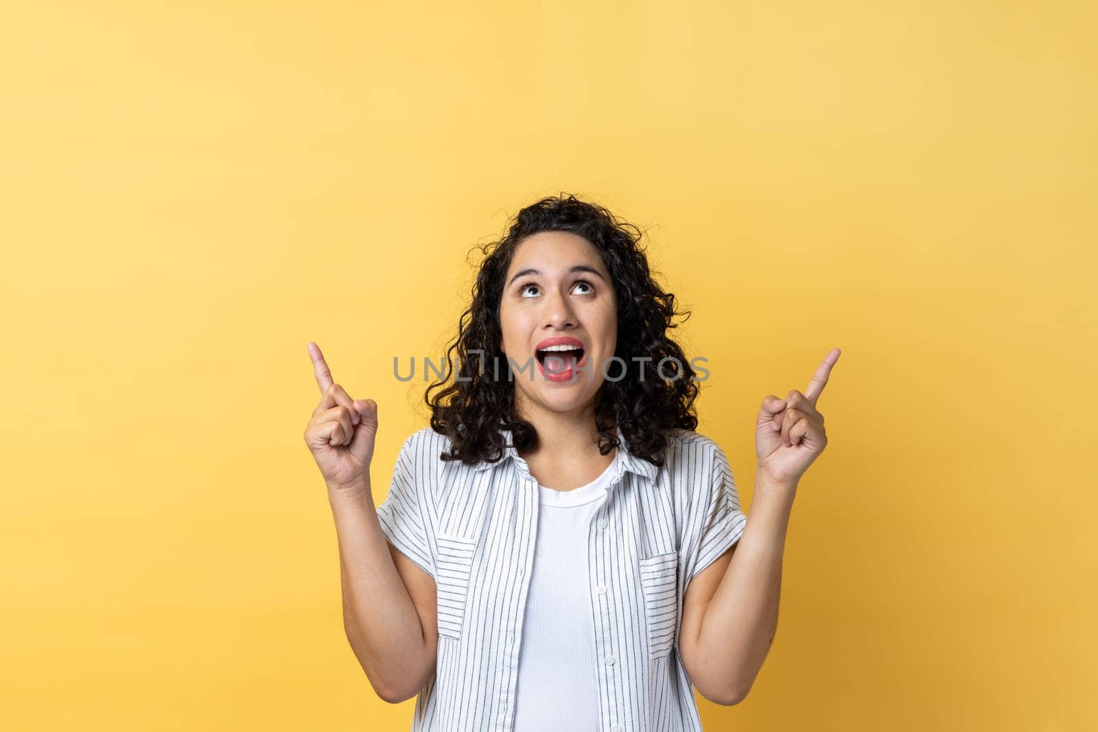 Wow, look, crazy advertisement. Portrait of woman with dark wavy hair pointing up at empty place for ad content and expressing astonishment. Indoor studio shot isolated on yellow background.