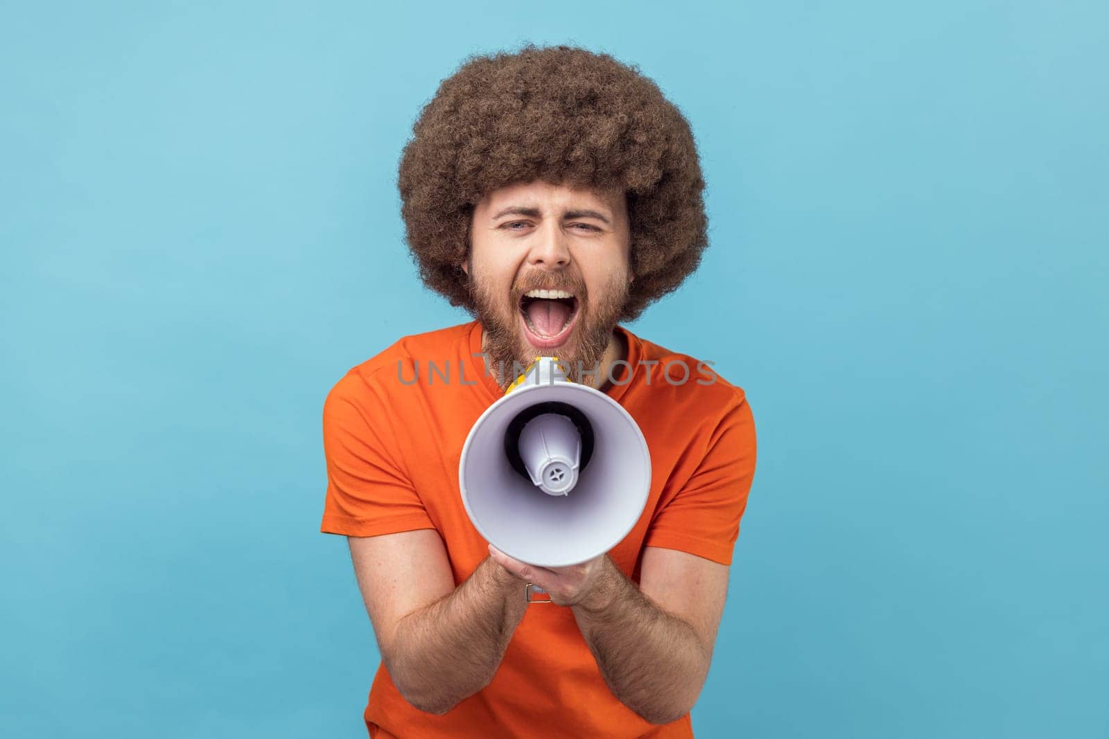 Portrait of man with Afro hairstyle wearing orange T-shirt loudly screaming at megaphone, making announce, protesting, wants to be heard. Indoor studio shot isolated on blue background.