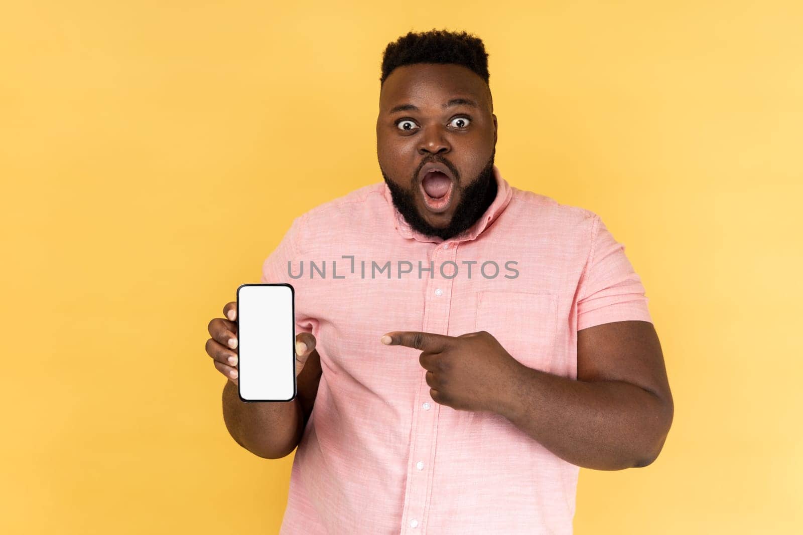 Portrait of shocked man wearing pink shirt pointing at cell phone screen, looking at camera with big eyes, showing mobile phone with white display. Indoor studio shot isolated on yellow background.