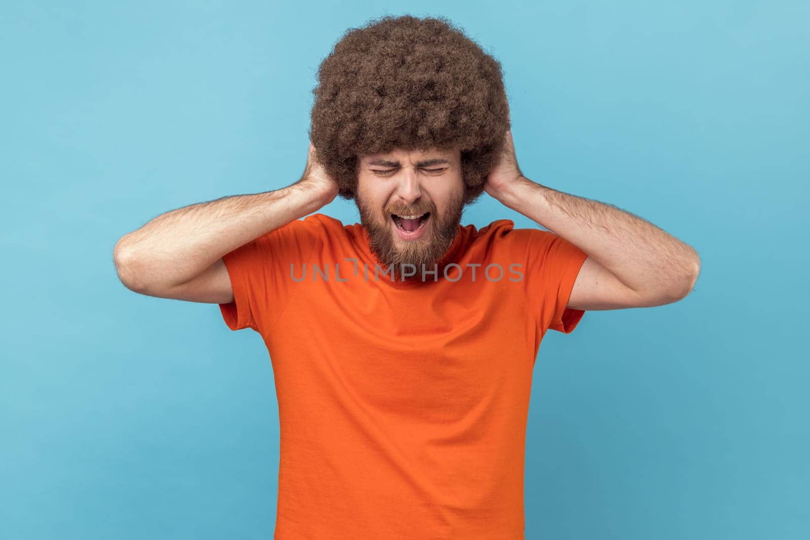 Portrait of man with Afro hairstyle covering ears, cannot focus in crowded place, feels discomfort over noise, being troubled to listen advice. Indoor studio shot isolated on blue background.