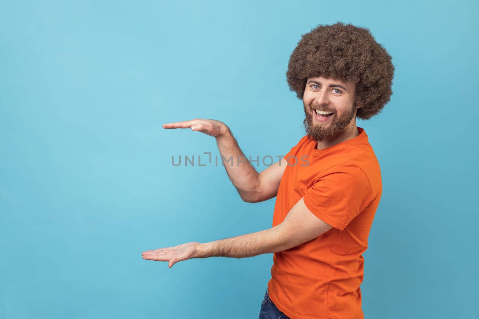 Portrait of positive optimistic man with Afro hairstyle presenting area for advertisement, looking at camera with toothy smile and optimism. Indoor studio shot isolated on blue background.