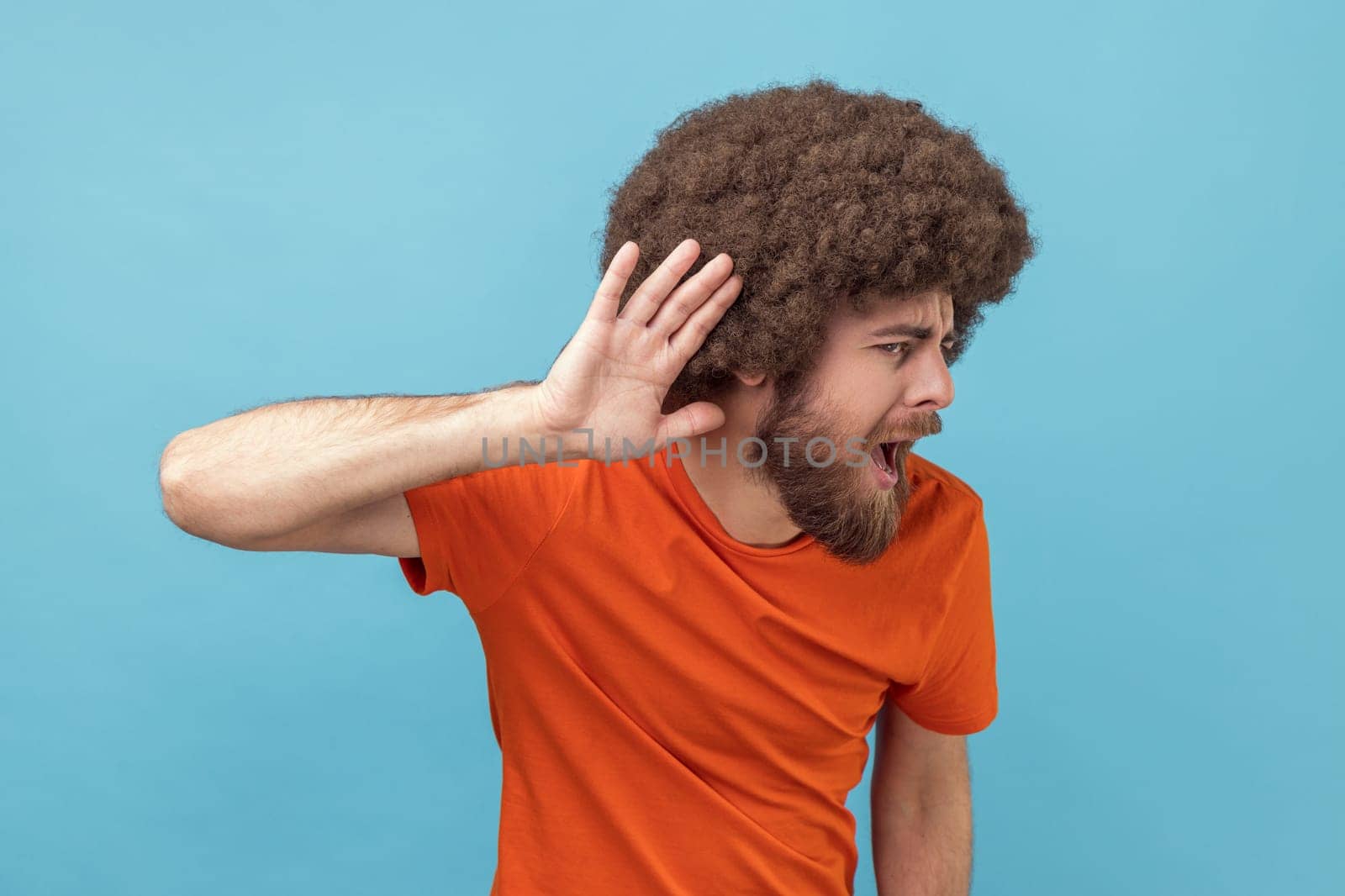 Portrait of man with Afro hairstyle in T-shirt holding hand near ear trying to listen quiet conversation, overhearing gossip, having hearing problems. Indoor studio shot isolated on blue background.