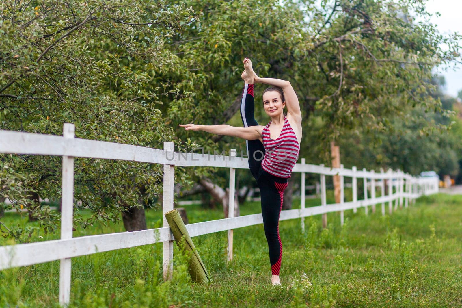 Full length portrait of flexible strong young girl stretching her leg, practicing yoga, having workout outdoor, raising leg in air, doing split stretch one foot, looking smiling away.