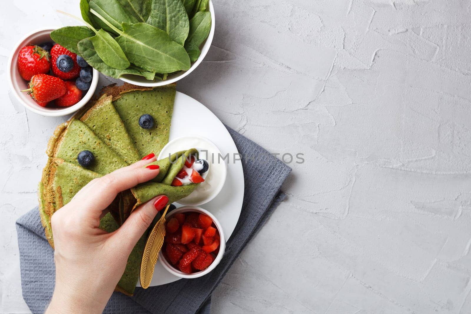 A woman's hand wets a spinach pancake with berries in a bowl with sour cream on a gray background with textiles. copy space.