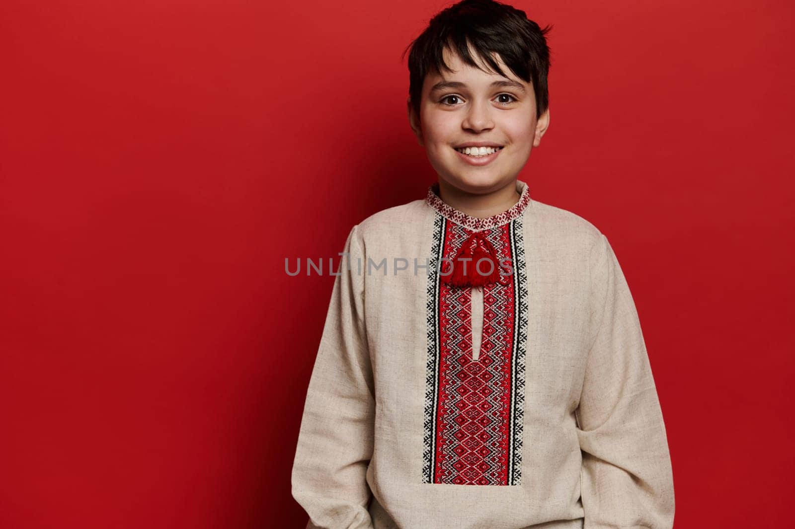 Portrait of a smiling Caucasian teen boy in Ukrainian traditional embroidered linen shirt, isolated on red background by artgf