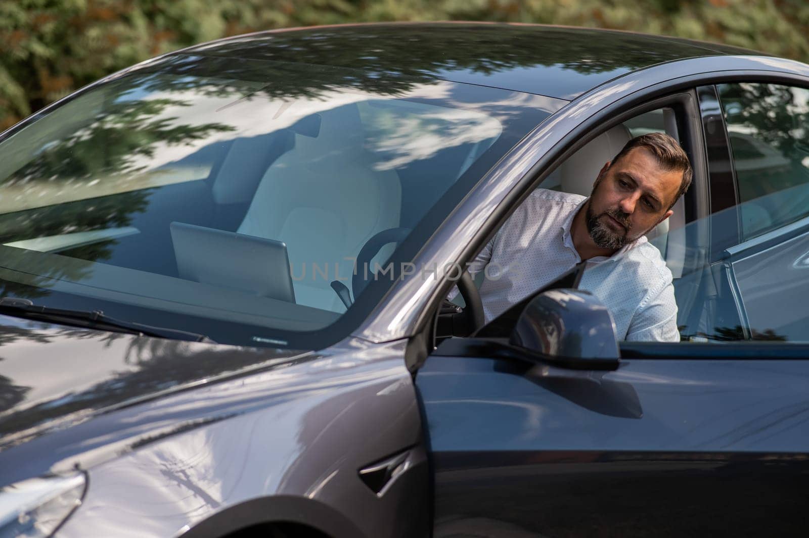 Caucasian bearded man in a suit gets out of a black electro car in the countryside in summer