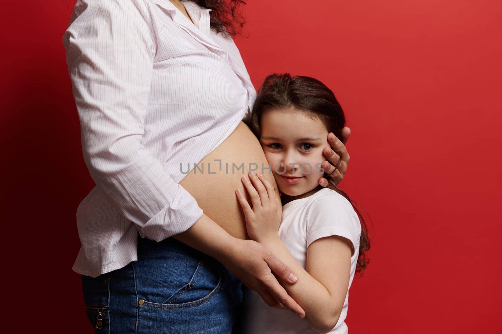 Beautiful little girl hugging the belly of her pregnant mother, looking at camera, isolated on red background. by artgf