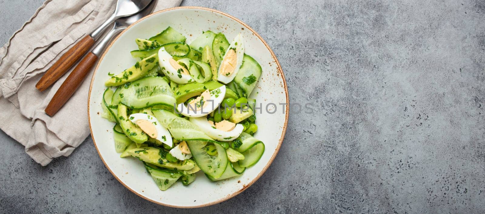 Healthy green avocado salad bowl with boiled eggs, sliced cucumbers, edamame beans, olive oil and herbs on ceramic plate top view, grey stone rustic table background. Copy space by its_al_dente