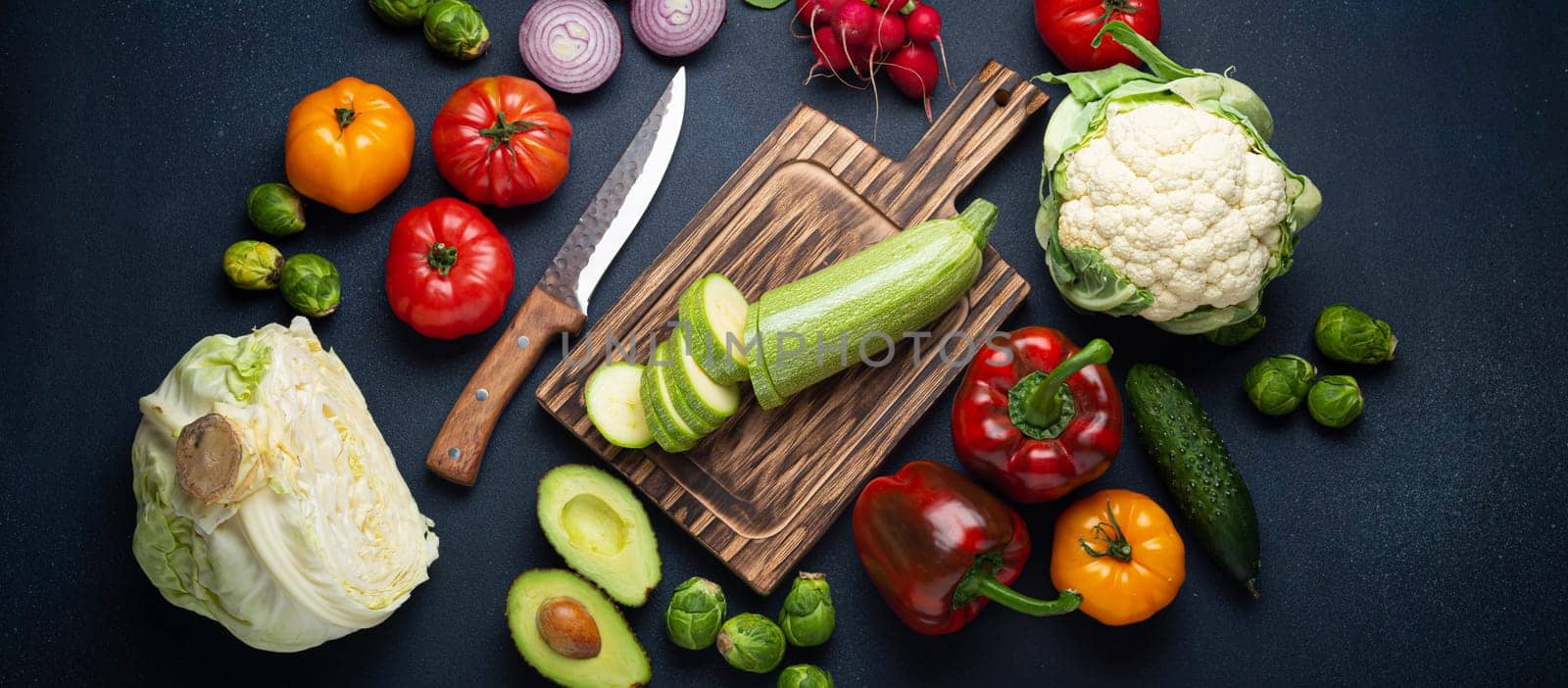 Fresh various vegetables, cut zucchini on wooden cutting board and knife on rustic dark background top view. Cooking vegetarian meal with healthy ingredients, diet food and nutrition concept by its_al_dente