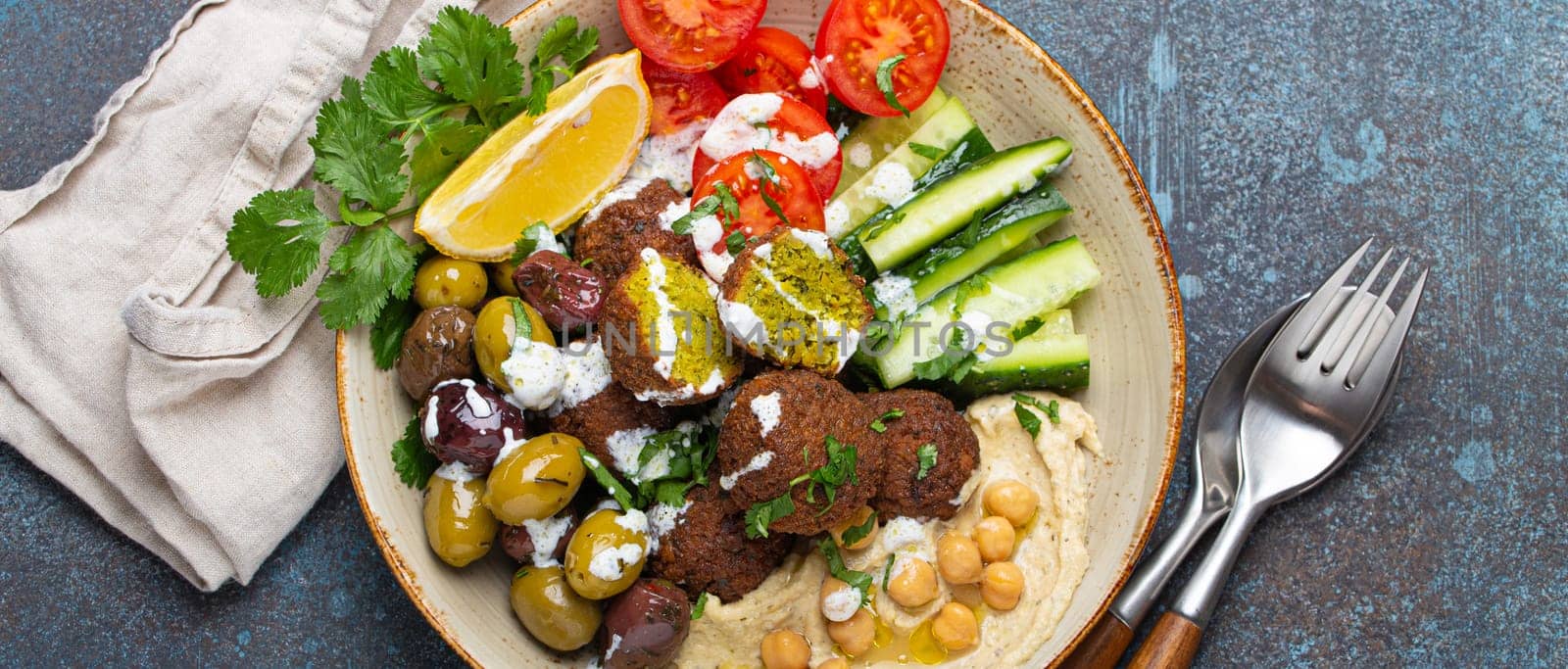 Falafel salad bowl with hummus, vegetables, olives, herbs and yogurt sauce. Vegan lunch plate top view on rustic stone background, healthy meal with falafel and veggies