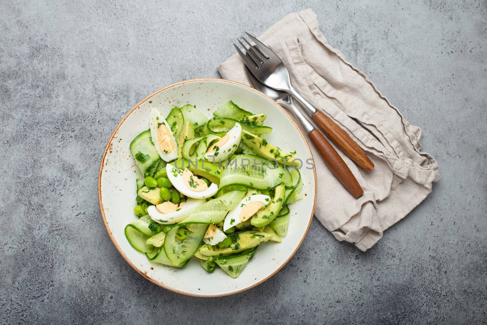 Healthy green avocado salad bowl with boiled eggs, sliced cucumbers, edamame beans, olive oil and herbs on ceramic plate top view on grey stone rustic table background