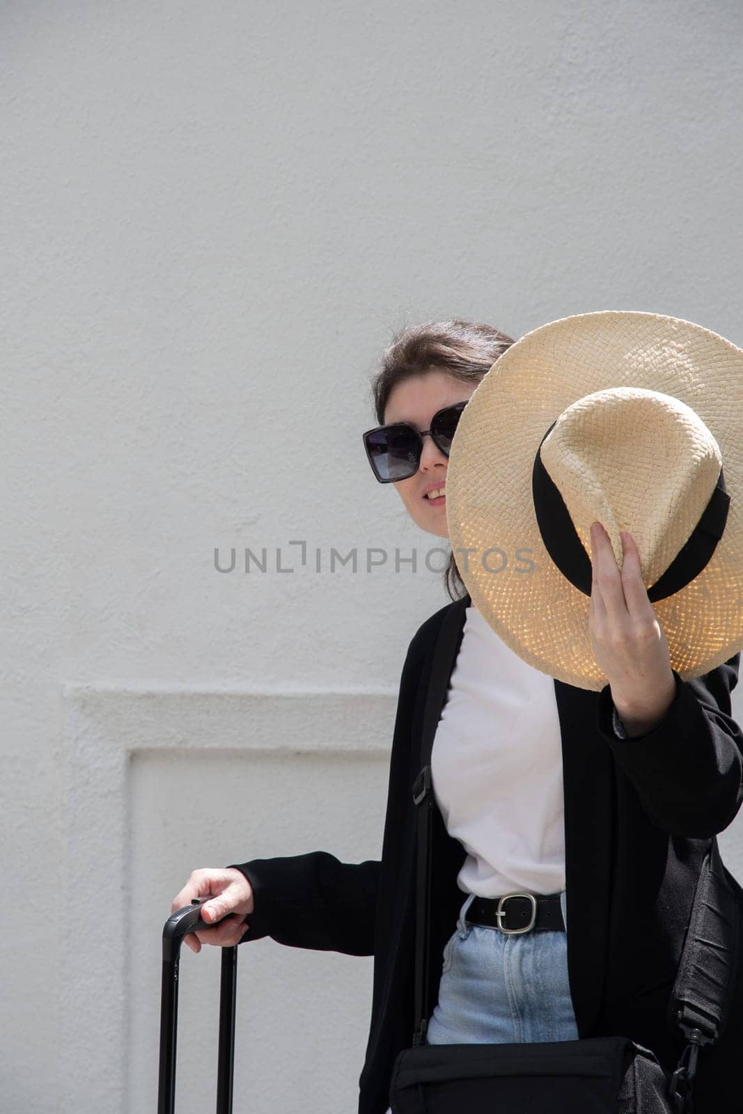 young happy brunette girl in sunglasses and straw hat with a suitcase goes on vacation, a woman stands against the background of a white brick wall, High quality photo