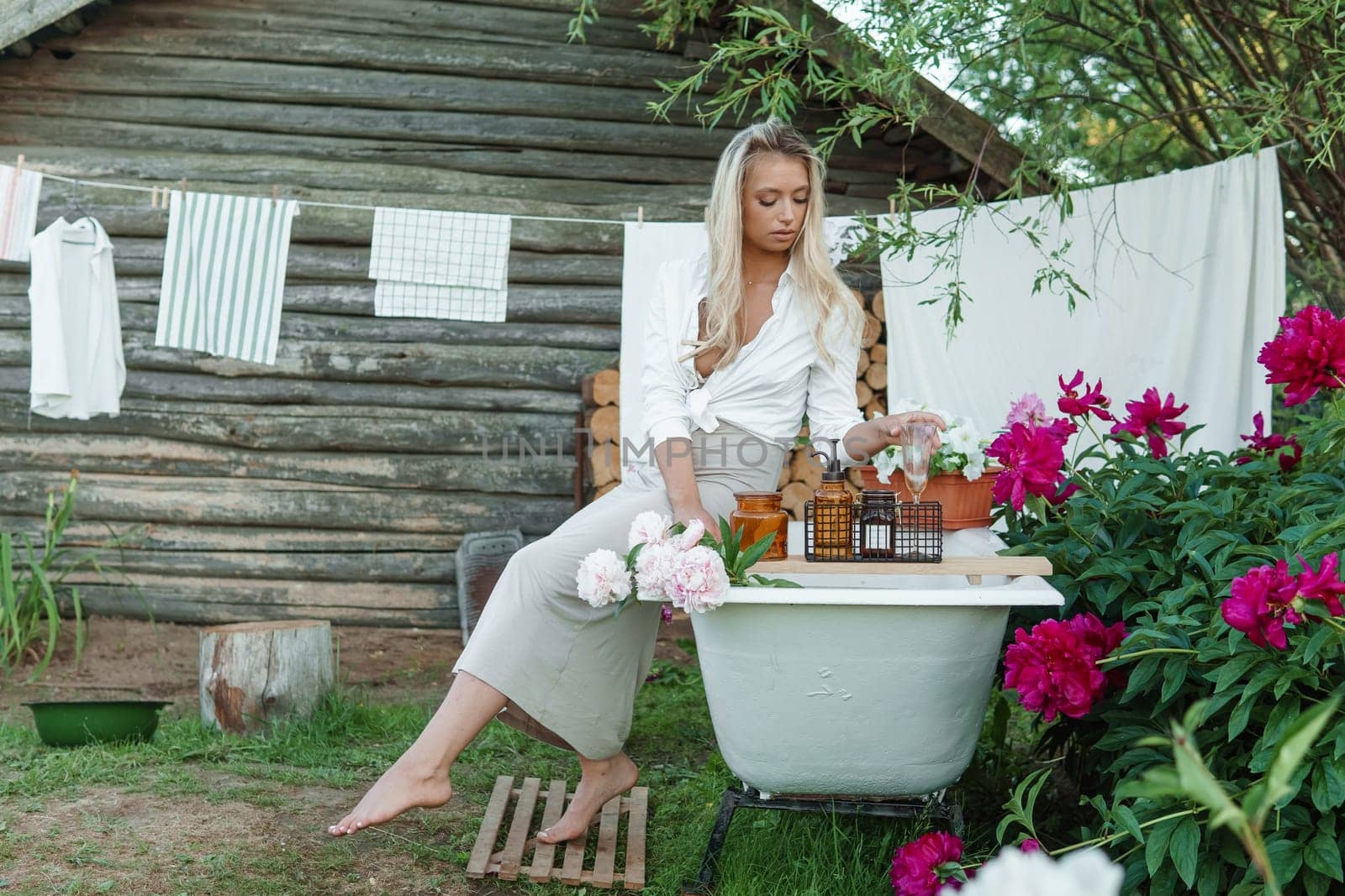 A woman is sitting on a cast-iron bathtub in the courtyard of a country house next to a bush of flowering peonies. The concept of summer, country life, a bathroom on the street in a blooming garden in the country