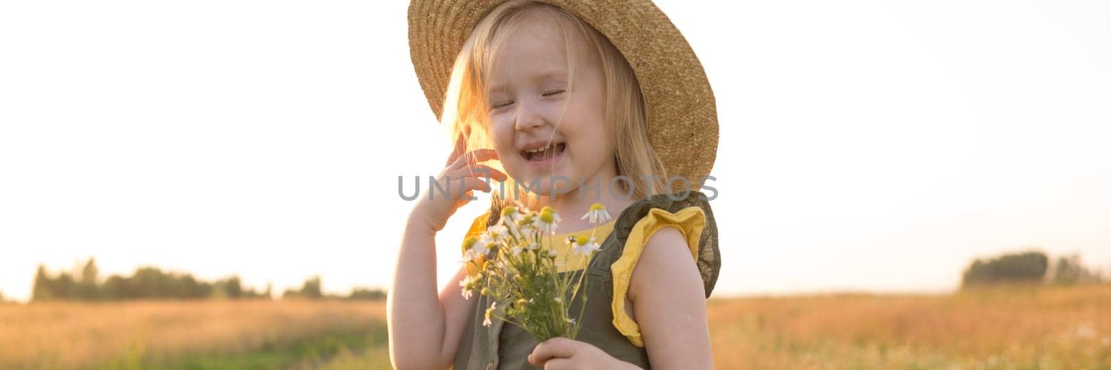A little blonde girl in a straw hat walks in a field with a bouquet of daisies. The concept of walking in nature, freedom and an eco-friendly lifestyle.