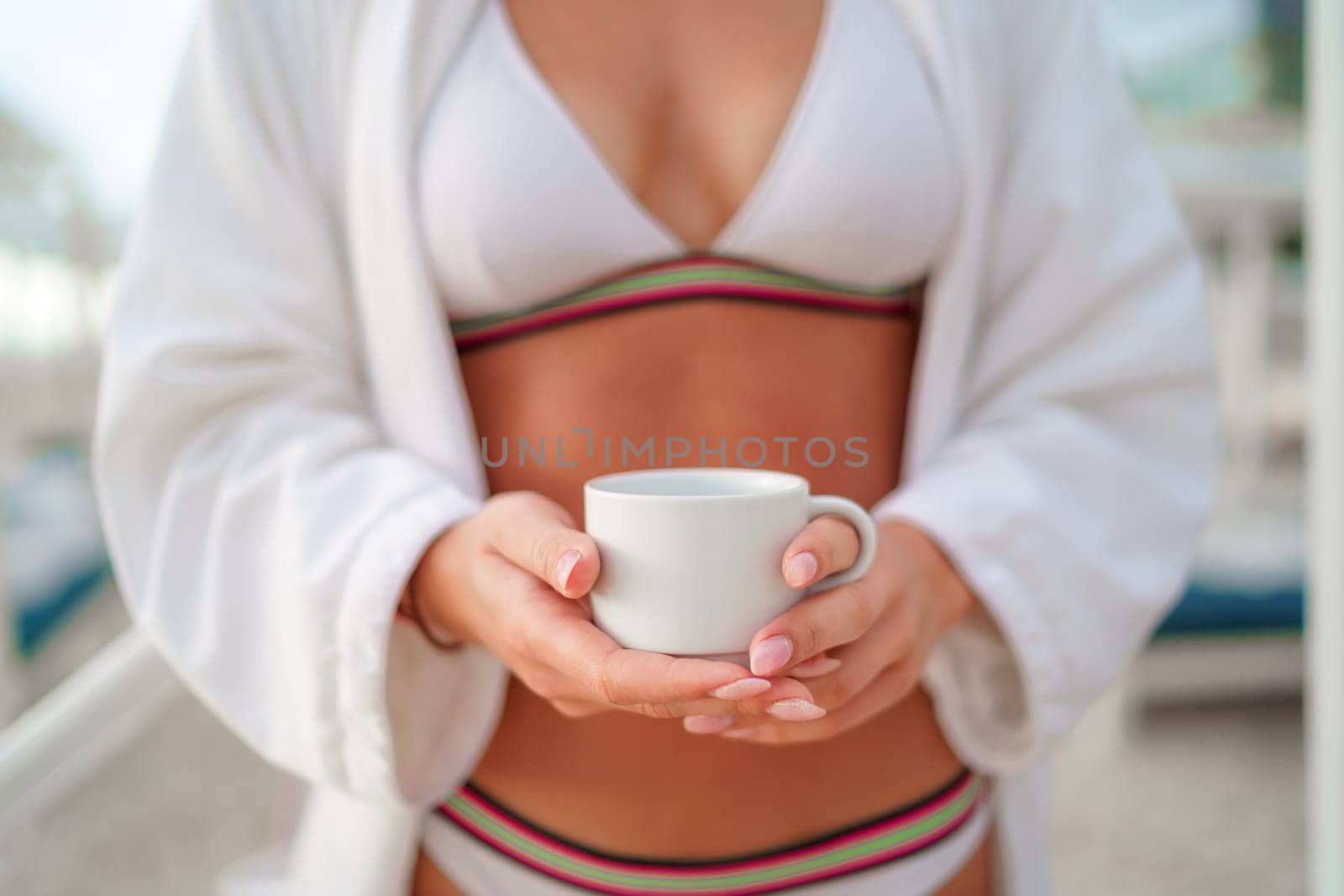 A white cup for coffee in the hands of woman in a bathing suit and a white bathrobe.