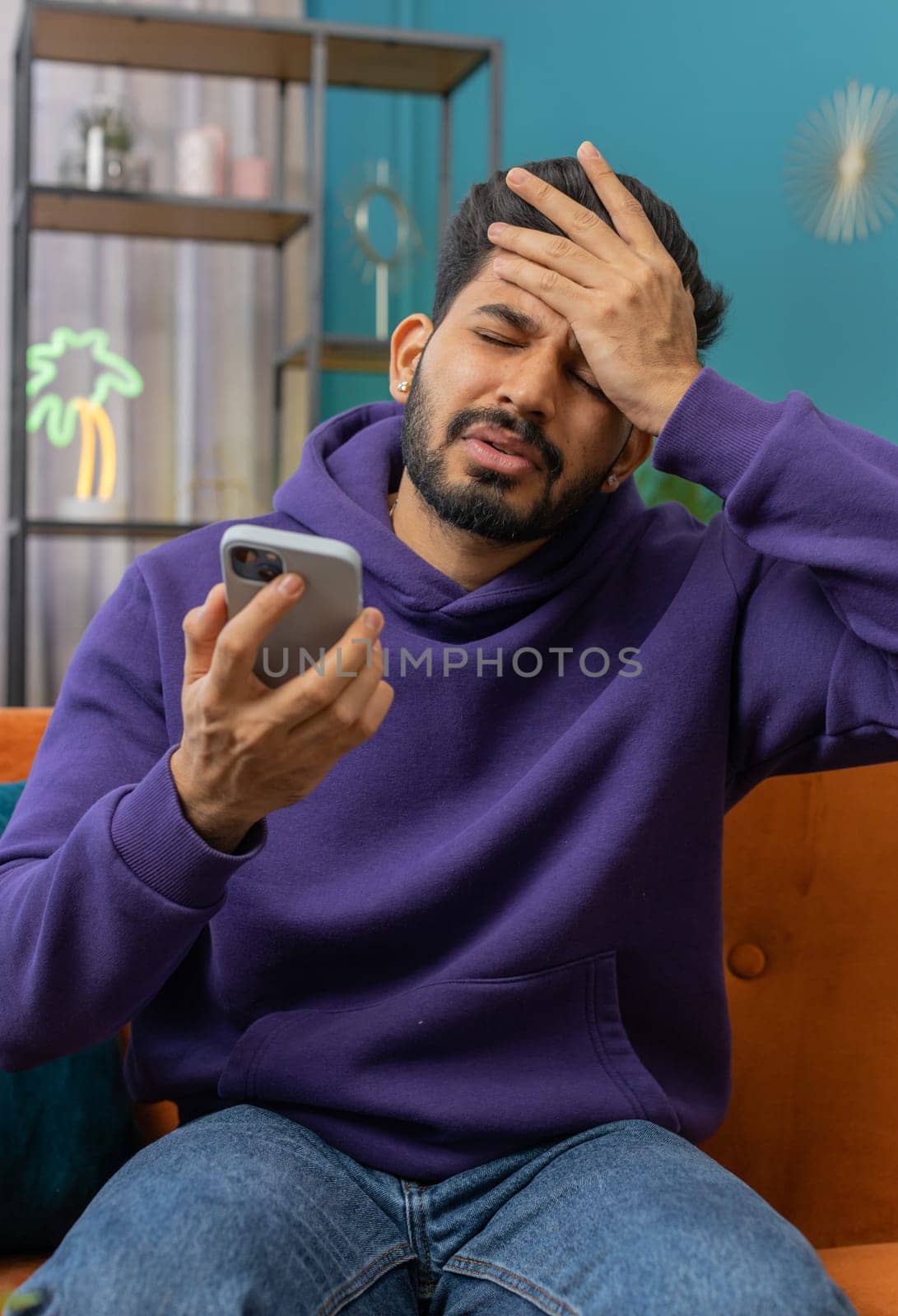 Sad displeased indian man use smartphone typing browsing, loses becoming surprised sudden lottery results, bad news, fortune loss, fail, deadline, virus. Young hindu guy at home living room on couch