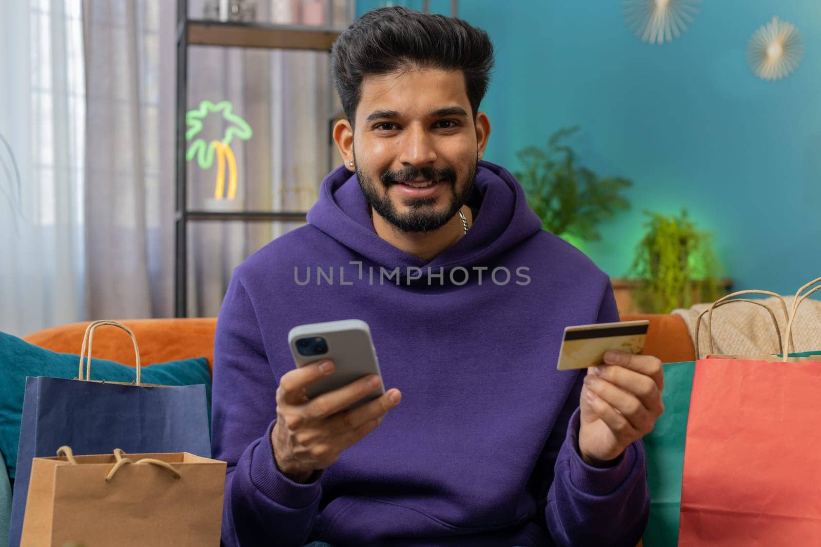 Happy shopaholic consumer indian man sitting with shopping bags at modern home apartment making online order, payment with credit plastic bank card. Discount sale on internet. Young hindu guy on sofa