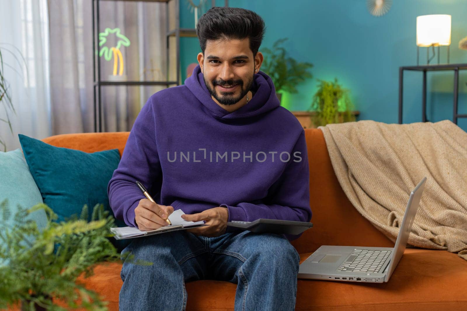 Indian man freelancer work with documents on home couch, reading information, business papers prepare financial report on laptop. Young hindu guy accountant analyzing graph documents, calculate taxes