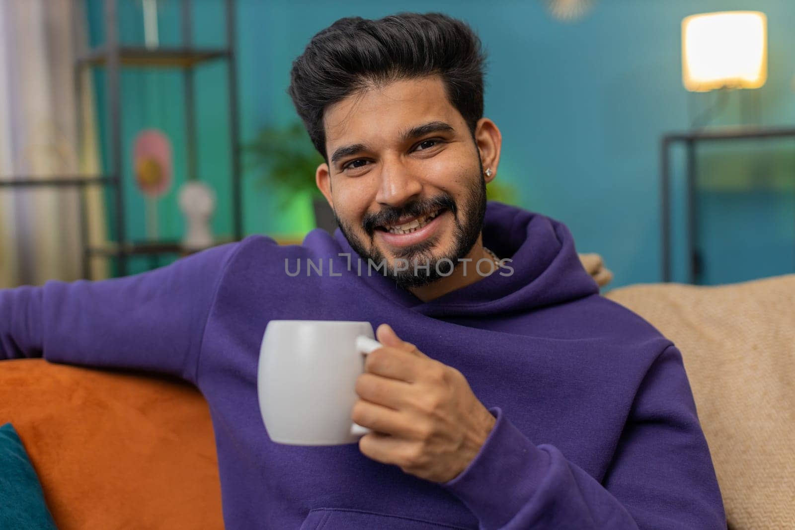 Handsome smiling indian man drinking a cup of warm coffee or herbal tea at home living room apartment. Break time. Hindu guy enjoying comfortable relaxing sitting on sofa in the morning. Lifestyles