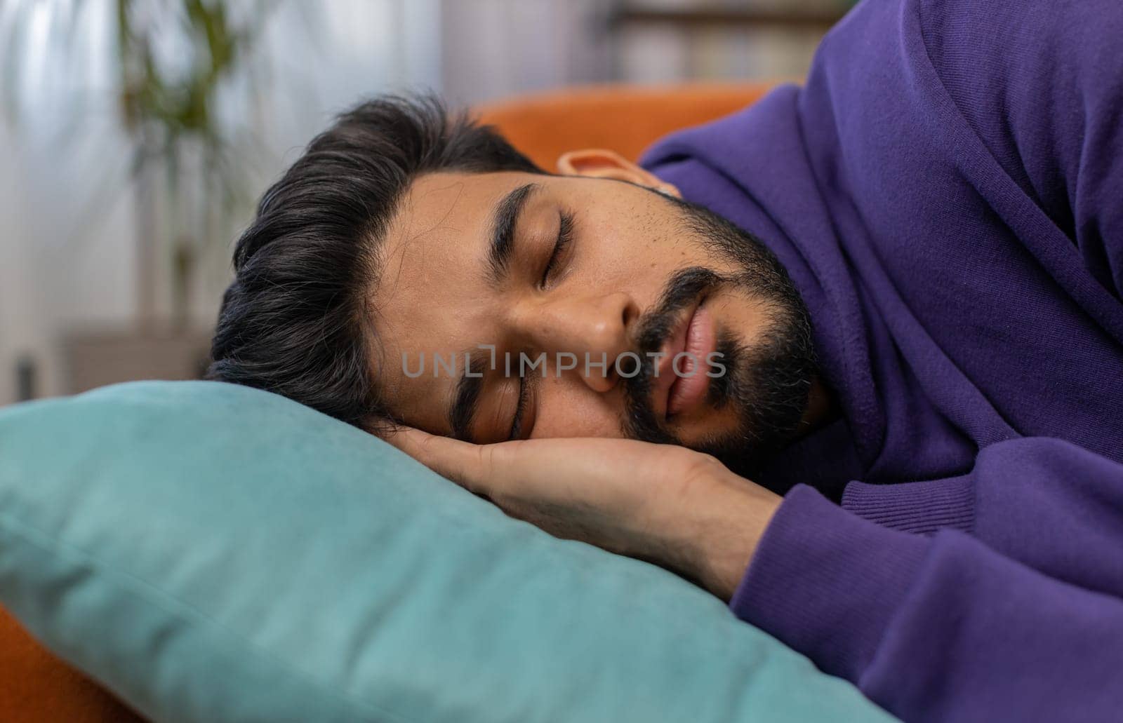 Tired indian man lying down in bed taking a rest. Carefree young hindu guy napping, falling asleep on comfortable sofa with pillows at home apartment room. Closed his eyes enjoy daytime nap alone