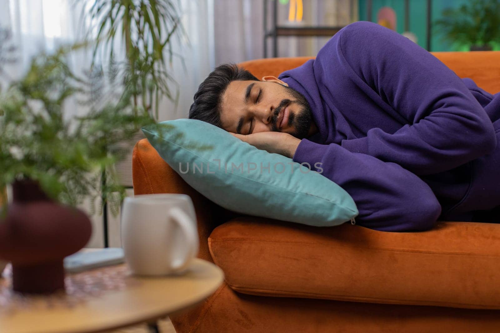 Tired indian man lying down in bed taking a rest. Carefree young hindu guy napping, falling asleep on comfortable sofa with pillows at home apartment room. Closed his eyes enjoy daytime nap alone
