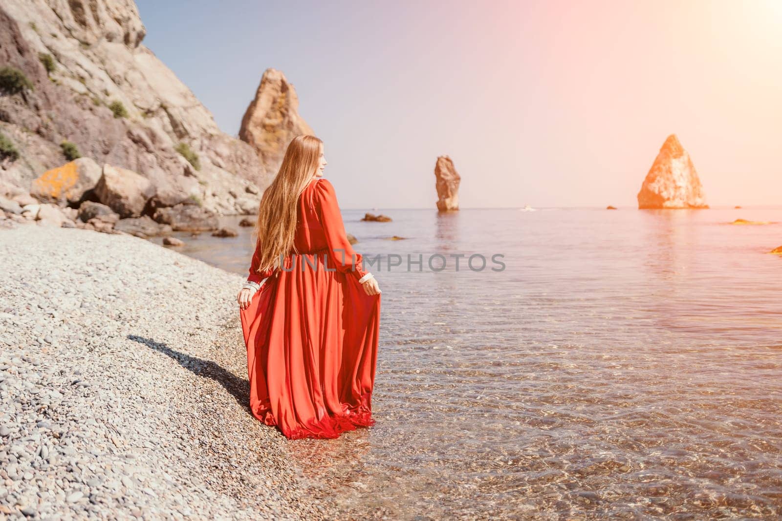 Woman travel sea. Happy tourist in red dress enjoy taking picture outdoors for memories. Woman traveler posing on the rock at sea bay surrounded by volcanic mountains, sharing travel adventure journey by panophotograph