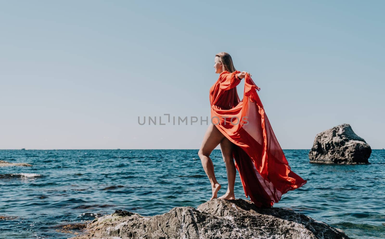 Woman travel sea. Young Happy woman in a long red dress posing on a beach near the sea on background of volcanic rocks, like in Iceland, sharing travel adventure journey