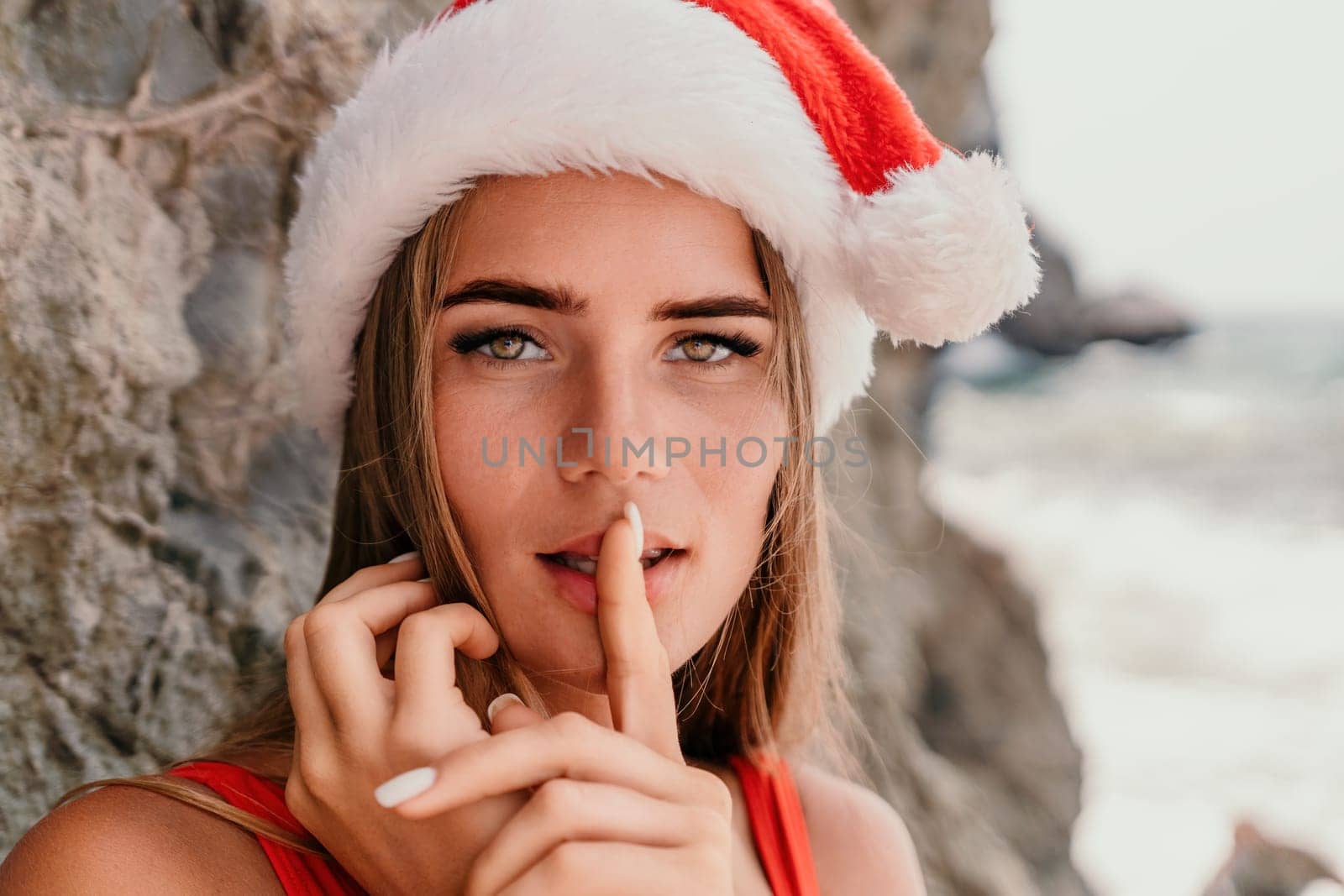 Woman summer travel sea. Happy tourist in red bikini and Santas hat enjoy taking picture outdoors for memories. Woman traveler posing on the beach surrounded by volcanic mountains, sharing travel joy by panophotograph