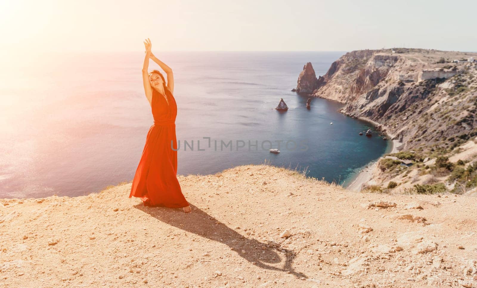 Side view a Young beautiful sensual woman in a red long dress posing on a rock high above the sea during sunrise. Girl on the nature on blue sky background. Fashion photo.