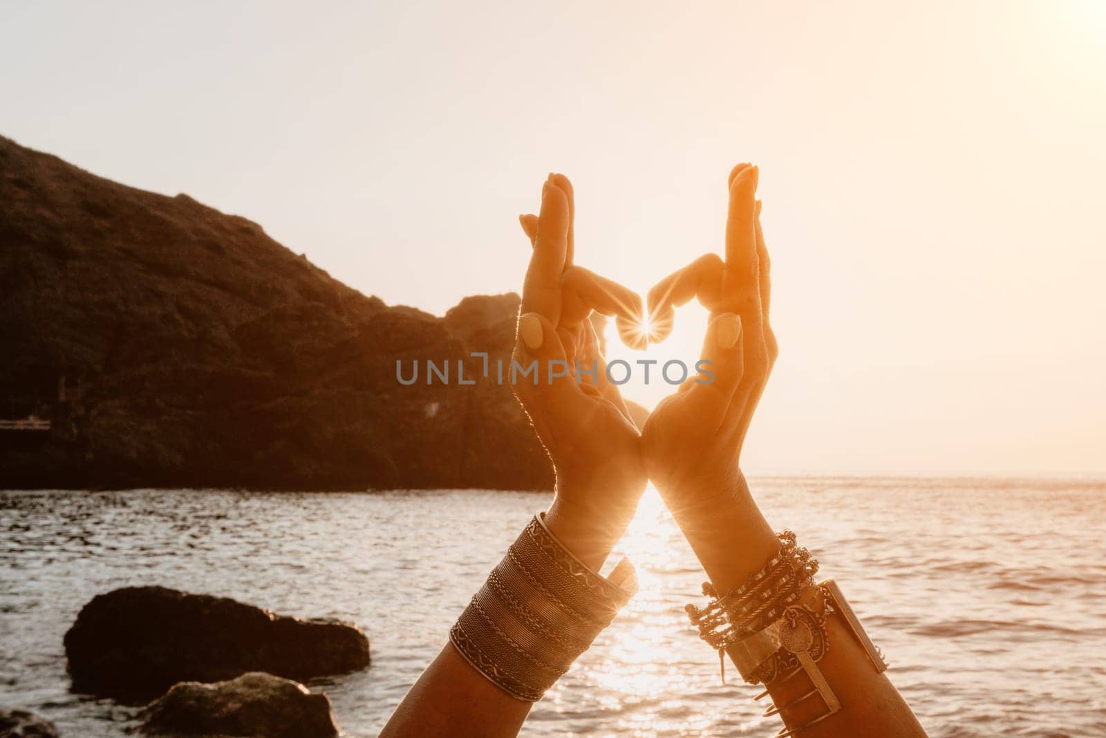 Woman sea yoga. Happy woman in a white swimsuit and boho-style bracelets making a heart shape with her hands while practicing yoga on a mat by sea at sunset. Healthy lifestyle, harmony and meditation by panophotograph