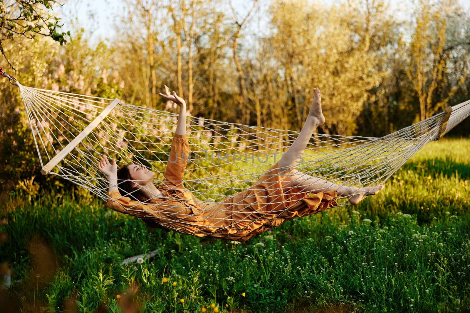 a funny woman is resting in nature lying in a mesh hammock in a long orange dress, lifting up her arms and legs, enjoying the rays of the setting sun by Vichizh