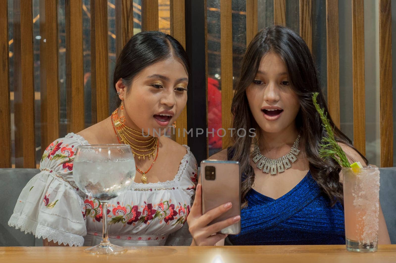 Multiracial friendship and cocktails in a modern restaurant while two girls are looking at a cell phone and being surprised. High quality photo