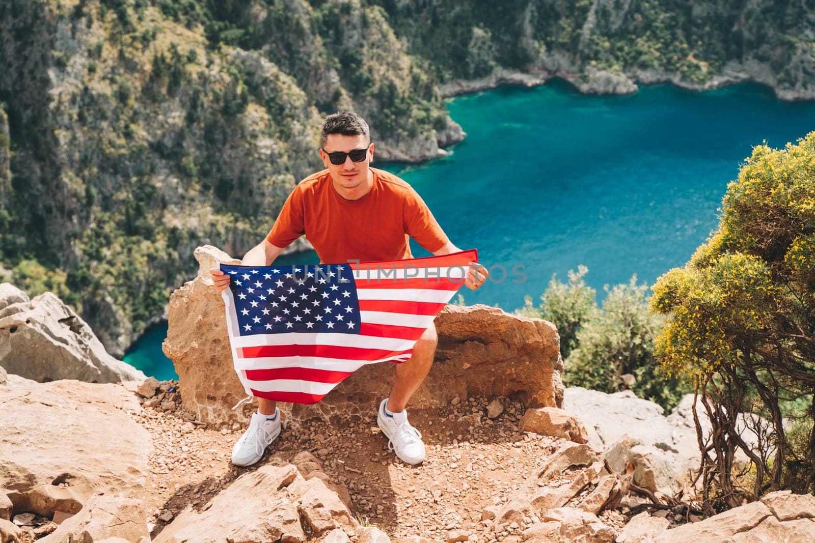 Young man sitting on a mountain top and holding the US flag in his hands. Veteran traveller holding the American flag while sitting on a mountain top. 4 fourth of July Independence Day by Ostanina