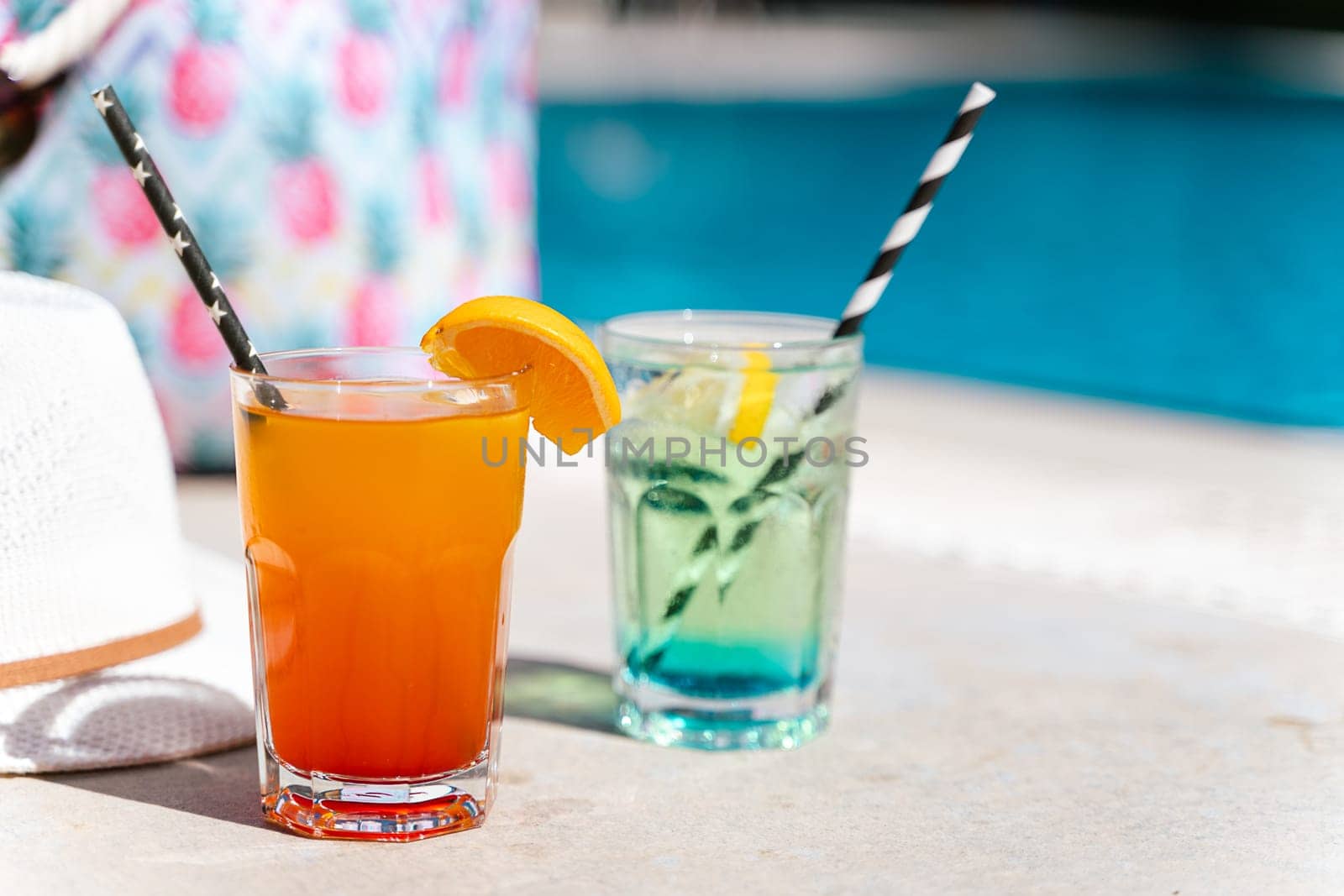 Tropical sparkling lemonade cocktails by the pool with pink beach bag and white hat in the background. Picture of glasses with orange and mint lemon fruit cocktails. Hello summer holiday vacation.