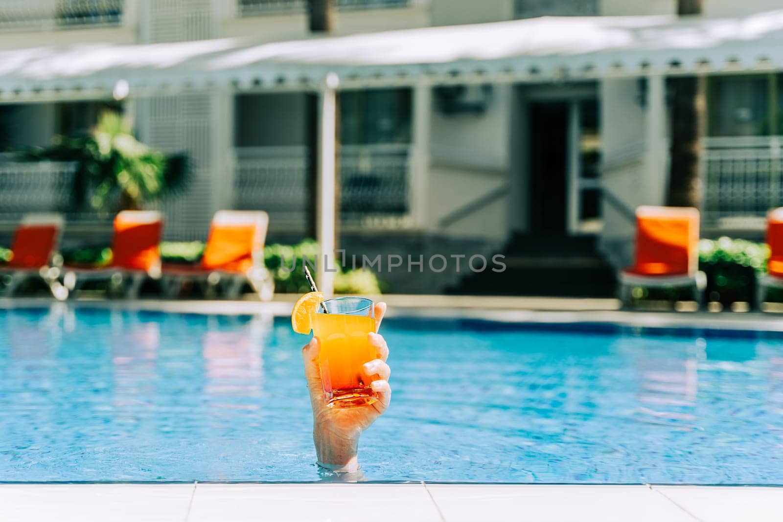 Closeup woman's hand sticking out of swimming pool holding tropical cocktail. Cropped girl's female's hand with an orange lemonade and the hotel pool in the background. Hello summer holiday vacation