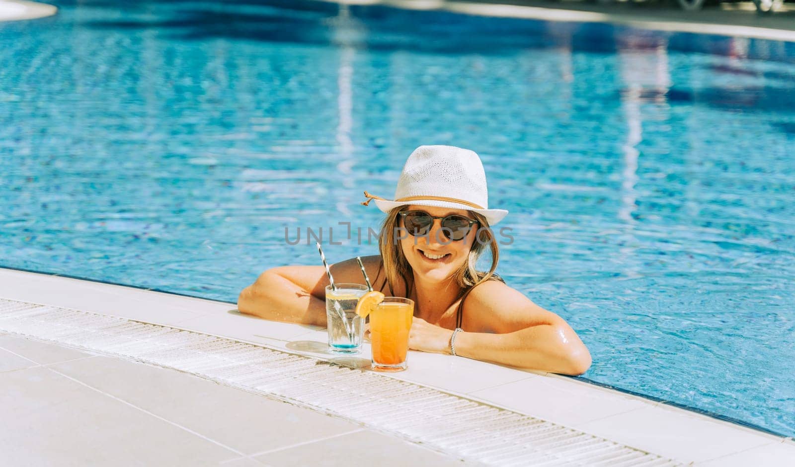 Portrait of beautiful cute smiling young woman lady girl in a bikini, hat, and sunglasses holding tropical lemonade cocktail while chilling in swimming pool. Hello summer holiday vacation